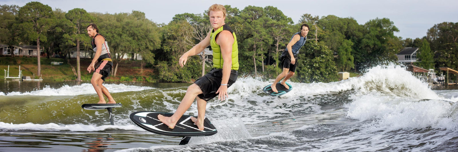 Three riders foiling across a lake with trees and homes in the background.