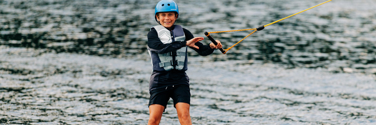 Kid riding a wakeboard in a vest and helmet while holding the tow rope on open water.