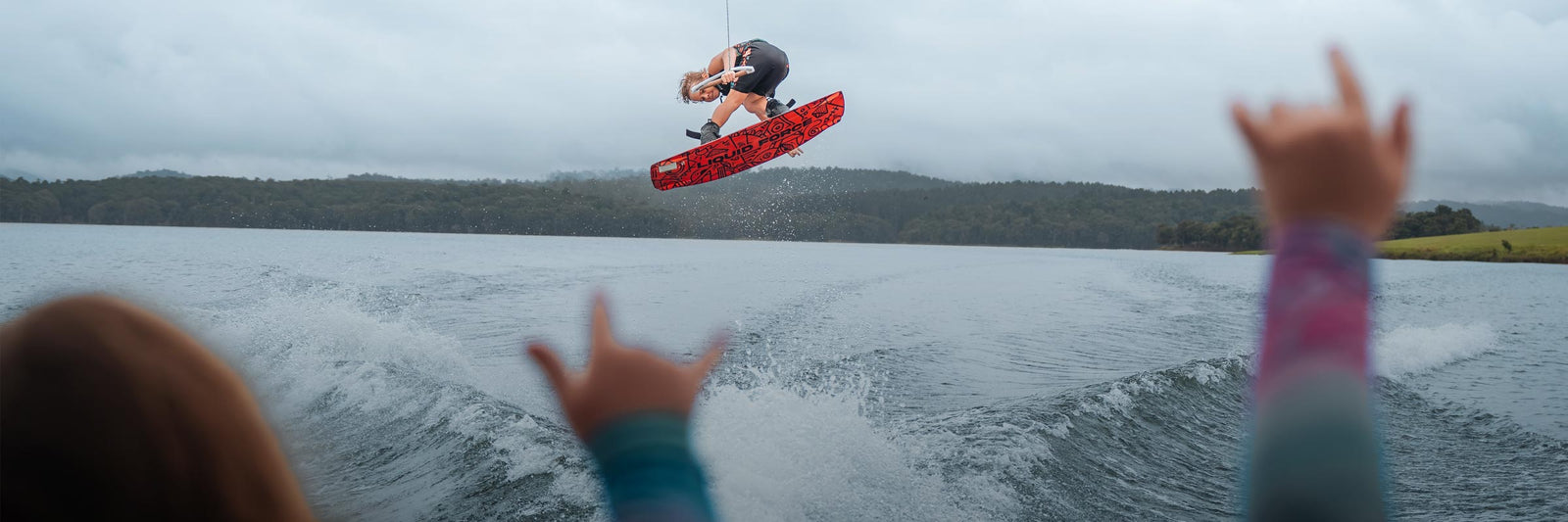 Young rider jumps a wake on a red board as two kids cheer from the boat in the foreground.