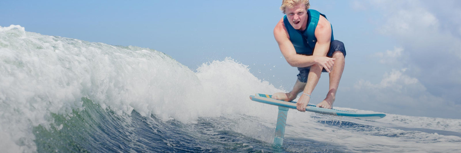 Rider crouches low on a foil board surfing a blue wave under a bright sky.