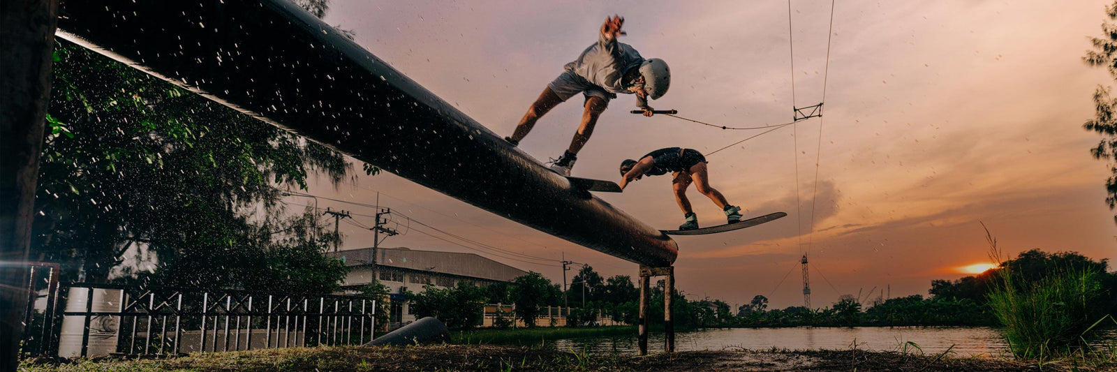 Two people wakeboarding on a large pipe against a sunset sky.