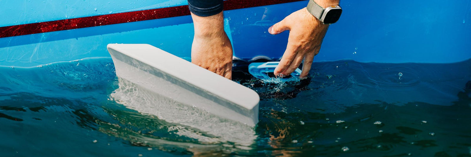 Hands place a wake shaper onto the side of a blue boat just above the waterline.