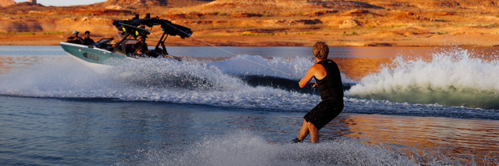Wakeboarder carving behind a boat on a scenic desert lake at sunset.