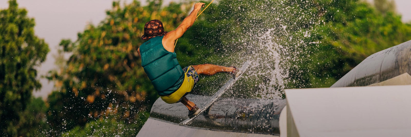 Rider grinds a rail on a wakeskate as water splashes against a backdrop of green trees.