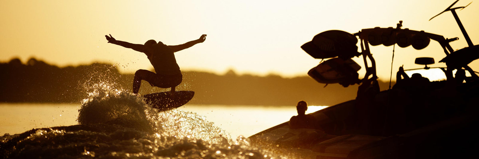 Silhouette of a wakesurfer catching air behind a boat at sunset with water spraying.