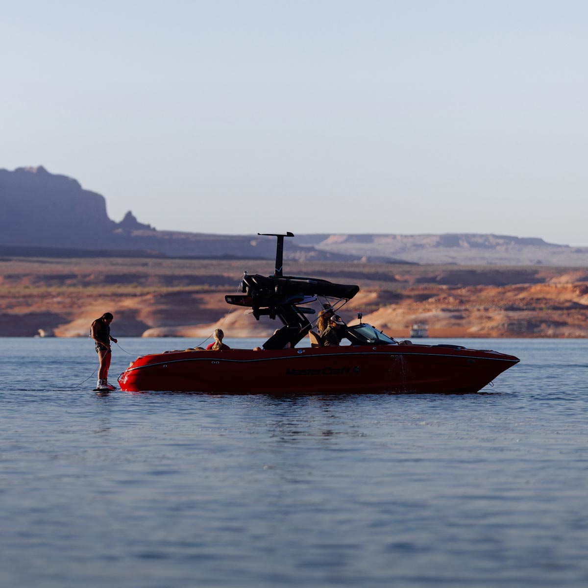 Rider standing beside a red boat on a calm lake with large desert rock formations behind.