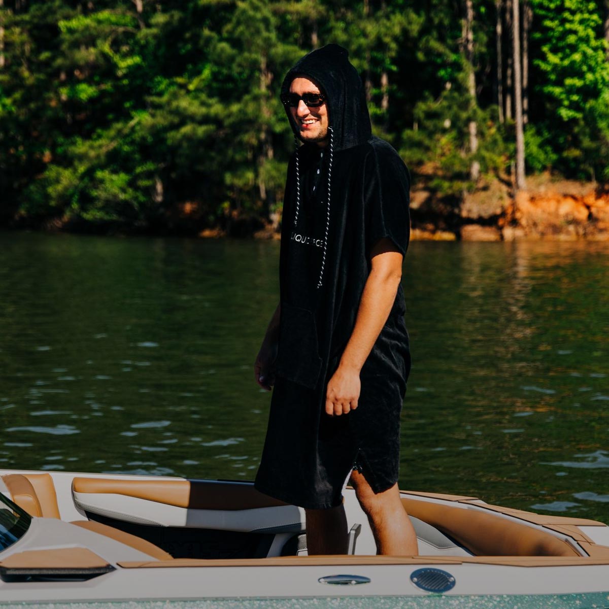 Man in a black changing poncho stands on a boat near a forested shoreline.