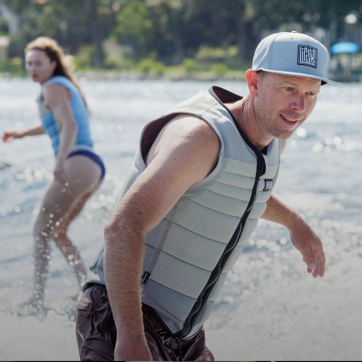 Riders foil surf on a lake, with one rider foregrounded wearing a branded hat.