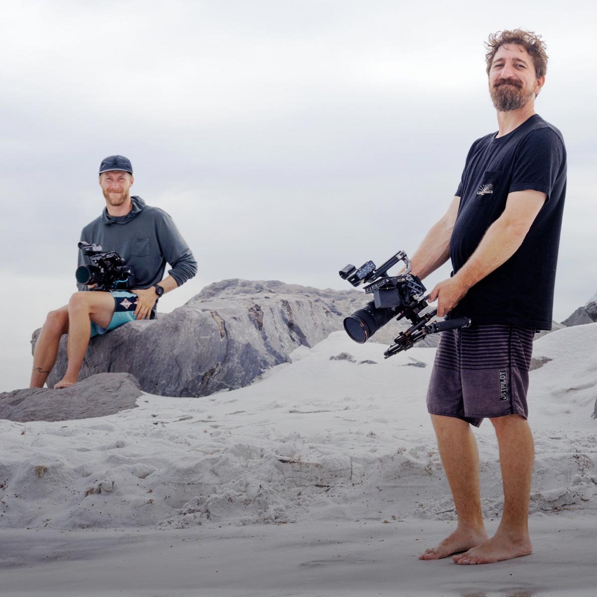 Two cameramen wearing Liquid Force shirts stand on rocky terrain with cameras.