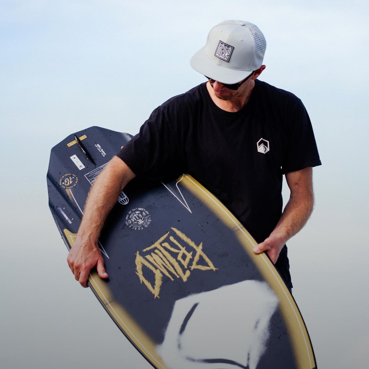 Man wearing a hat and black tee carries a wakesurf board by the water.
