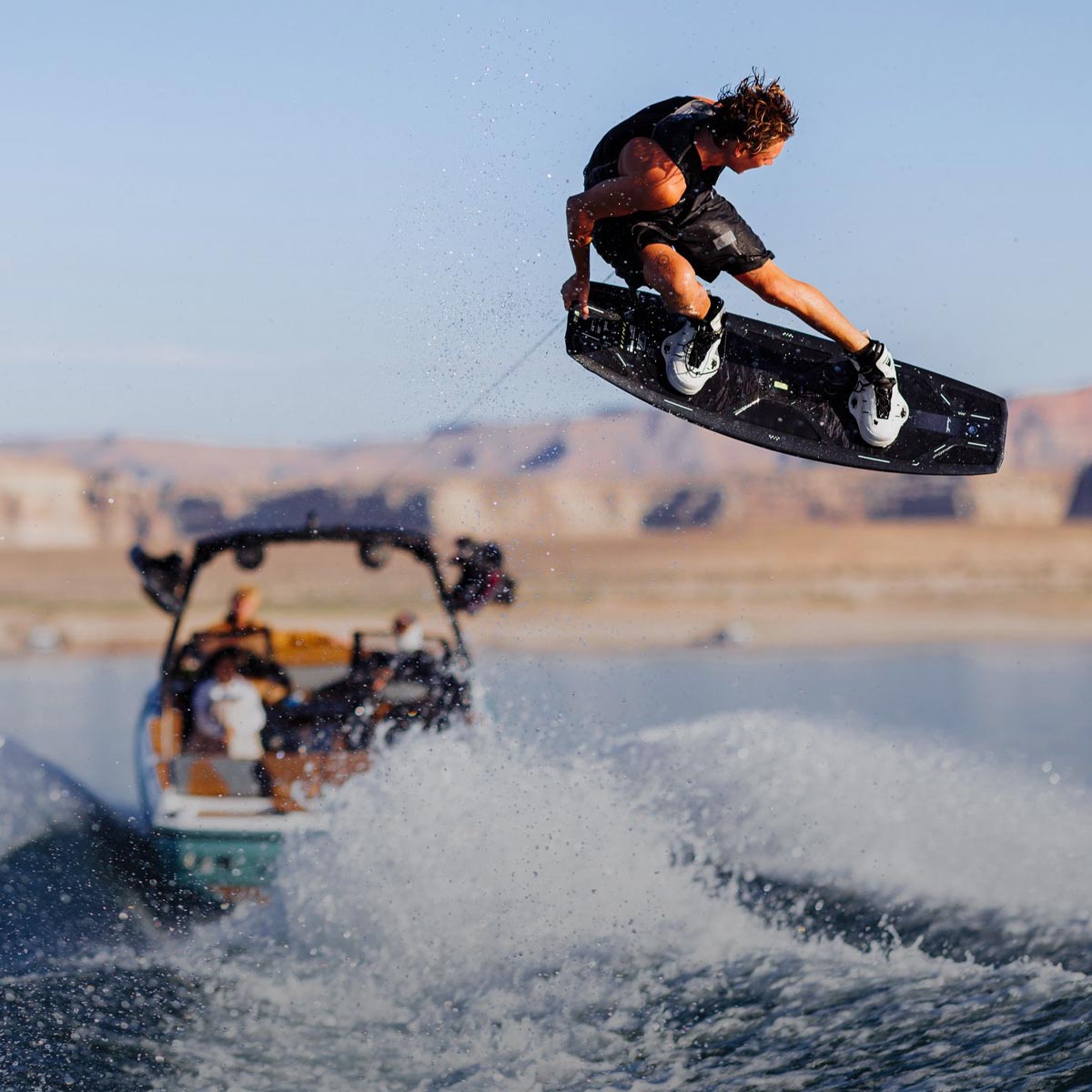 Rider launches off a wake behind a boat on a sunny lake with desert cliffs in the background.