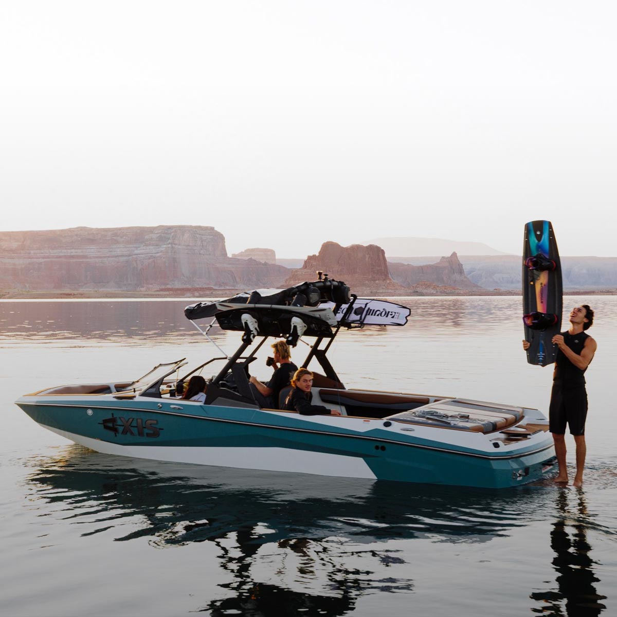 Rider holding a wakeboard beside a teal boat at sunset on a quiet lake.