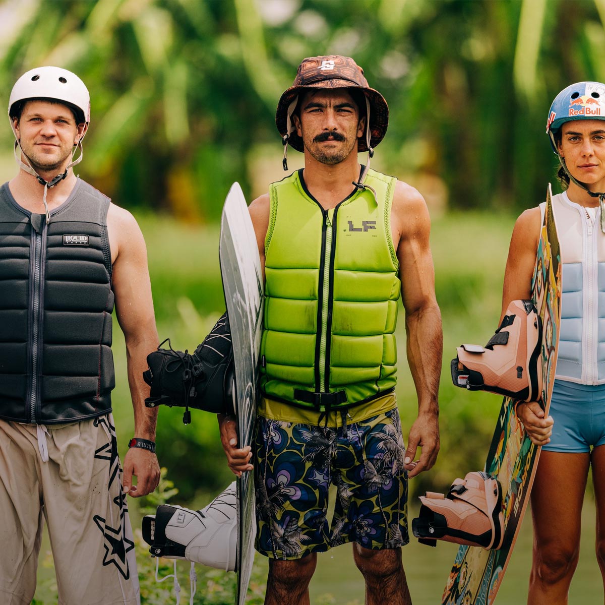 Three riders in comp vests and helmets stand side by side holding wakeboards.