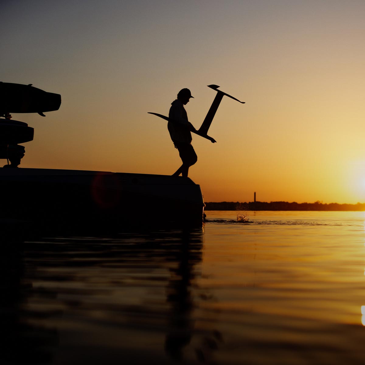 Silhouette of a rider carrying a foil board beside a boat during a calm orange sunset.