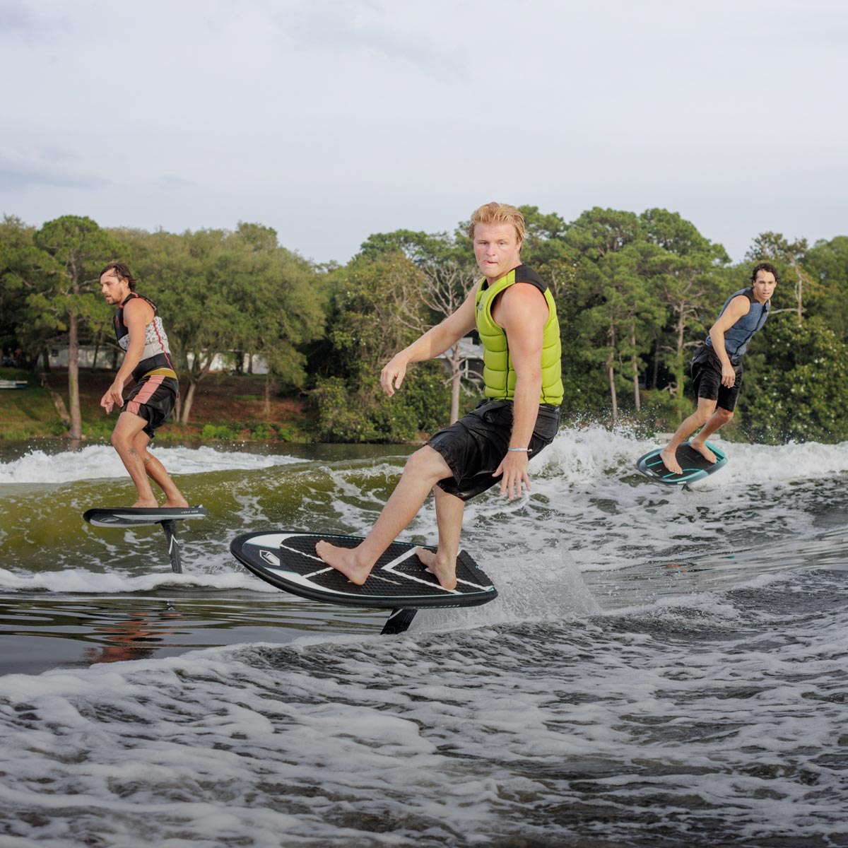 Three riders foiling across a lake with trees and homes in the background.