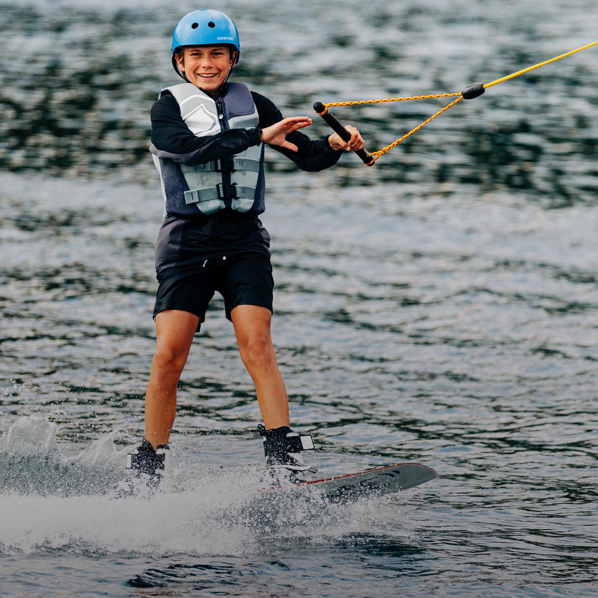 Kid riding a wakeboard in a vest and helmet while holding the tow rope on open water.