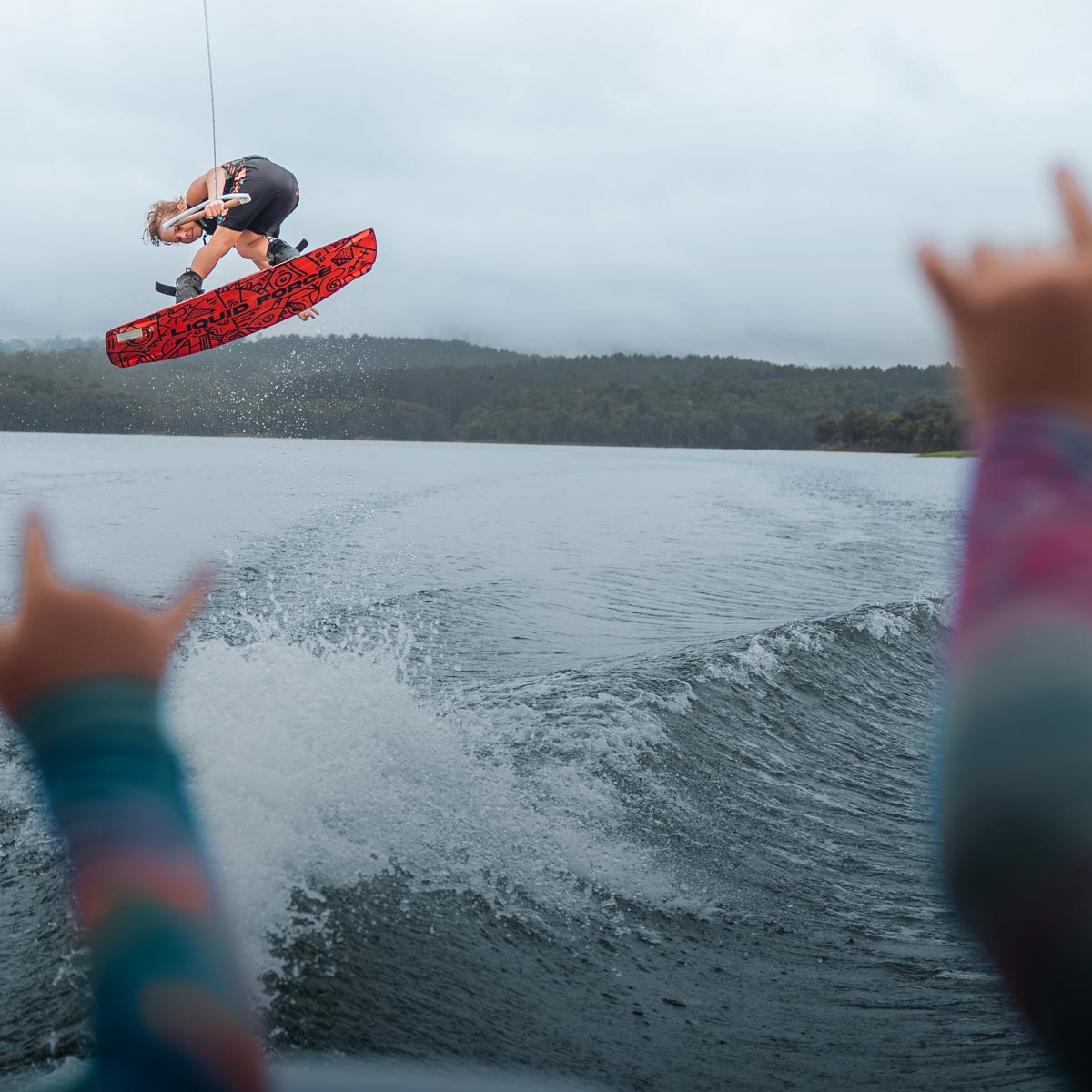Young rider jumps a wake on a red board as two kids cheer from the boat in the foreground.
