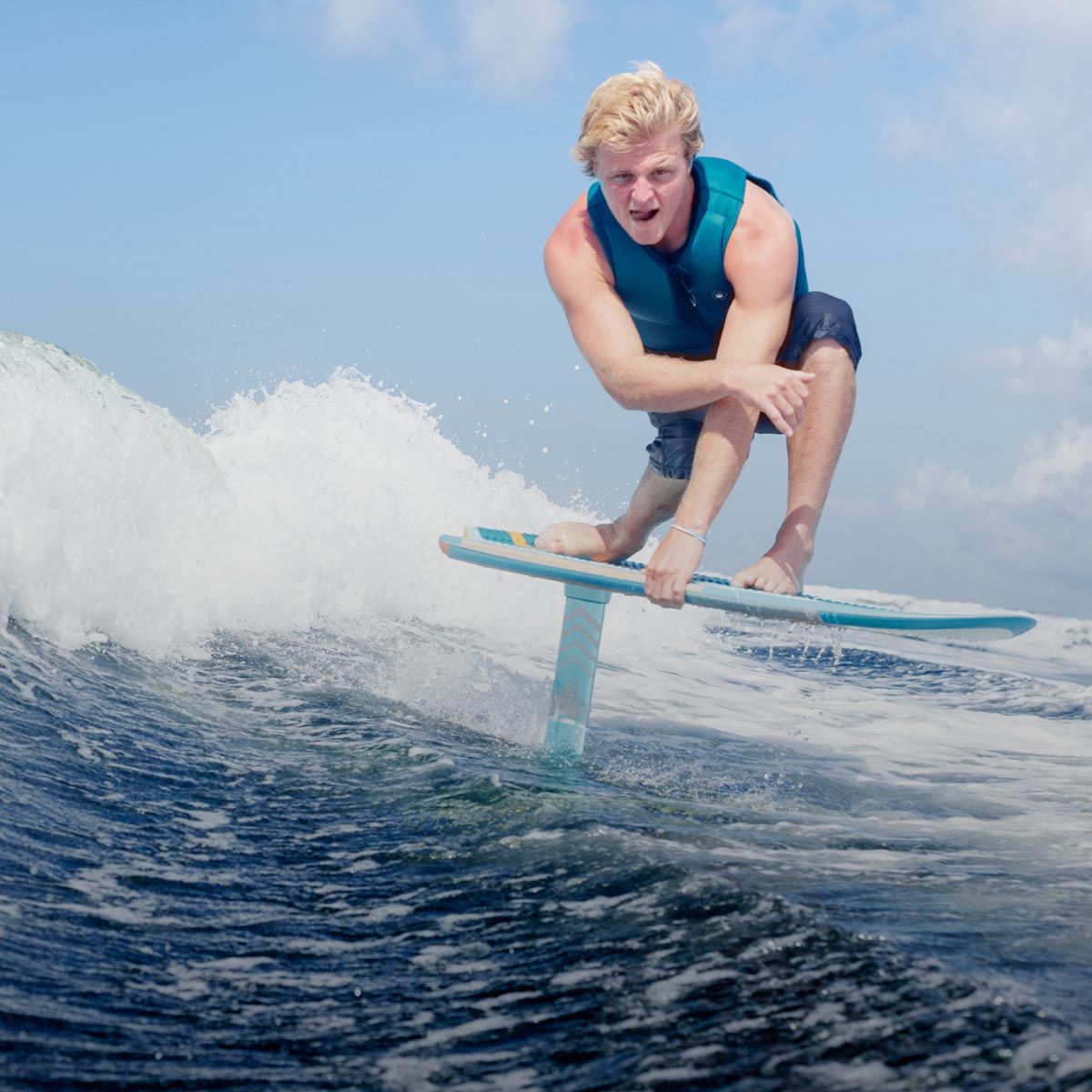 Rider crouches low on a foil board surfing a blue wave under a bright sky.