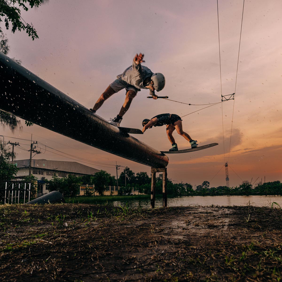 Two people wakeboarding on a large pipe against a sunset sky.