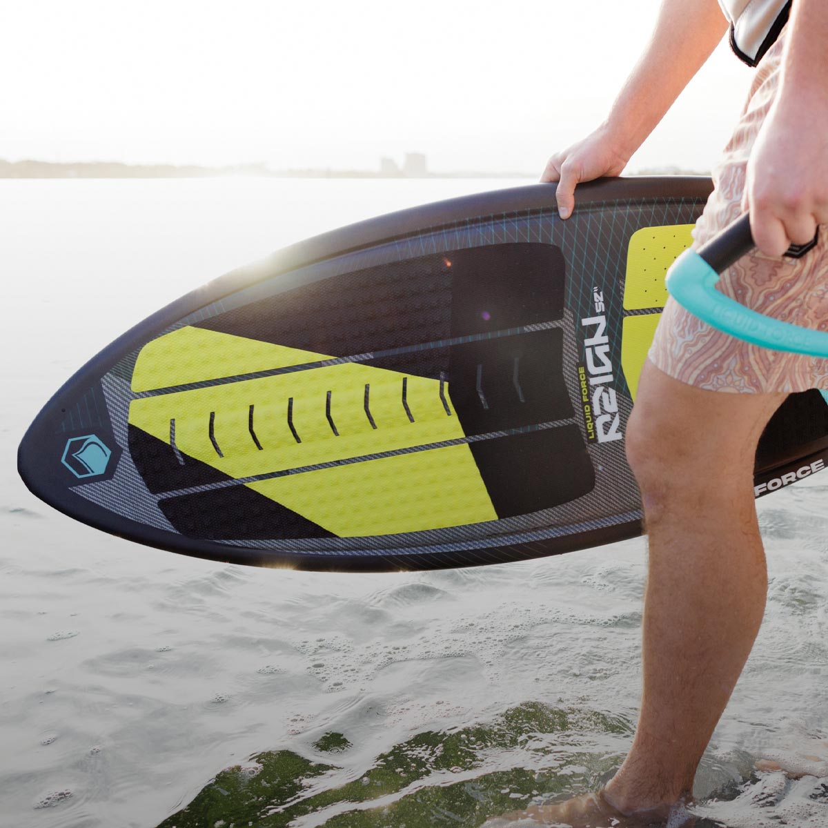 Rider holds a skim-style board and rope handle while standing ankle-deep in water.