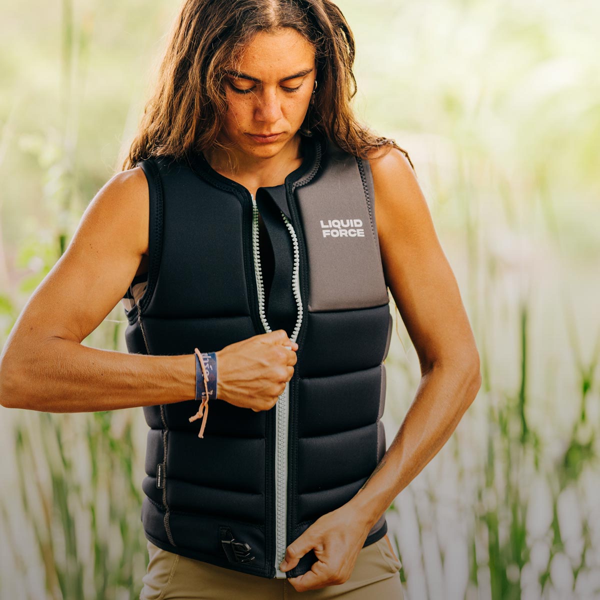 Woman zipping up a black padded life vest outdoors with soft greenery in the background.