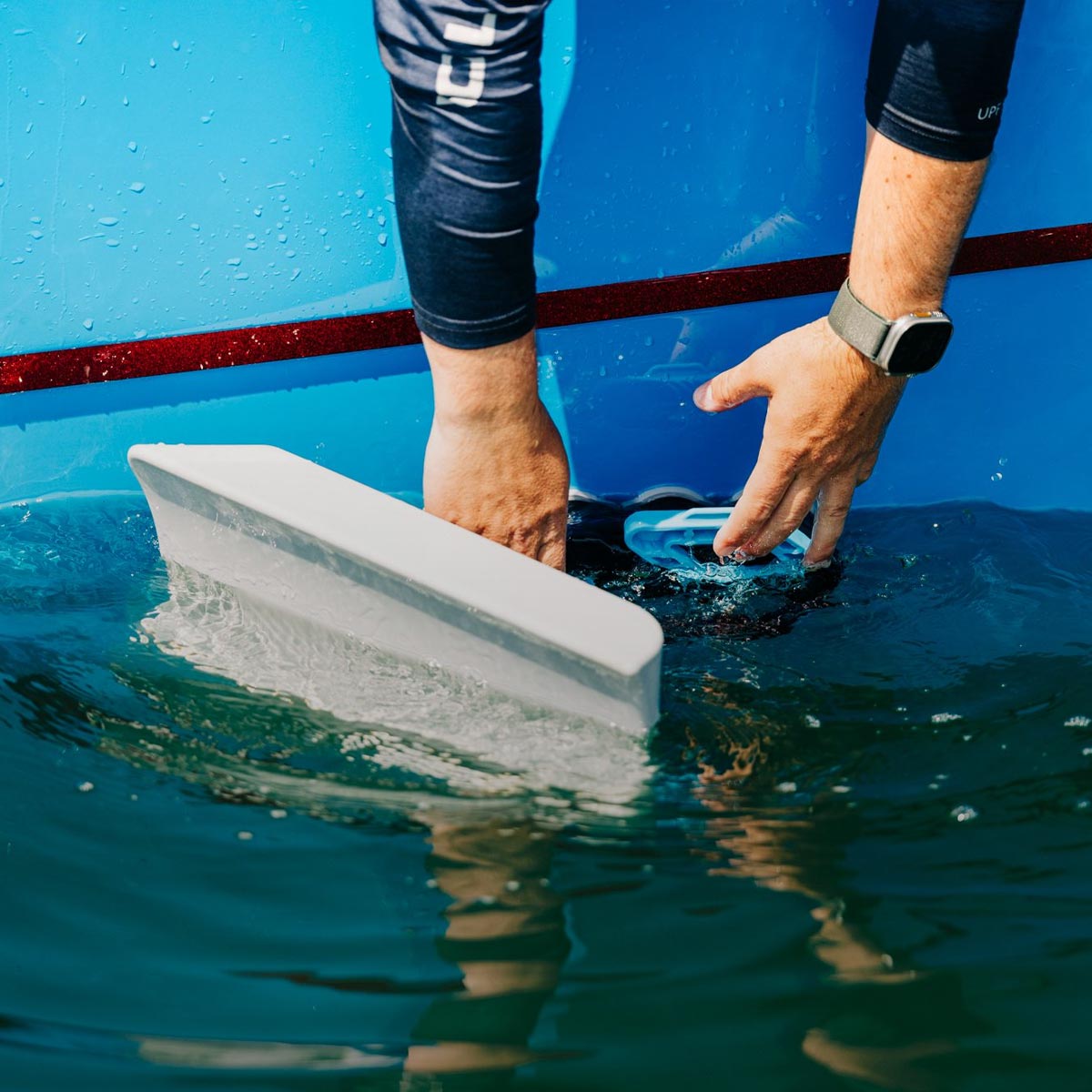 Hands place a wake shaper onto the side of a blue boat just above the waterline.