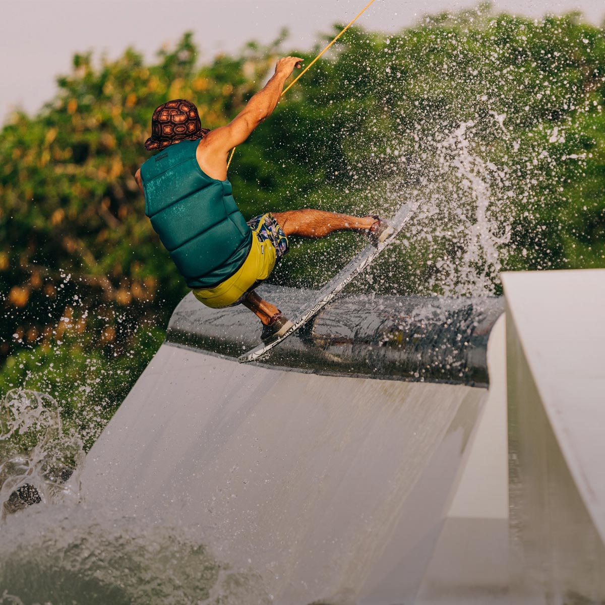 Rider grinds a rail on a wakeskate as water splashes against a backdrop of green trees.
