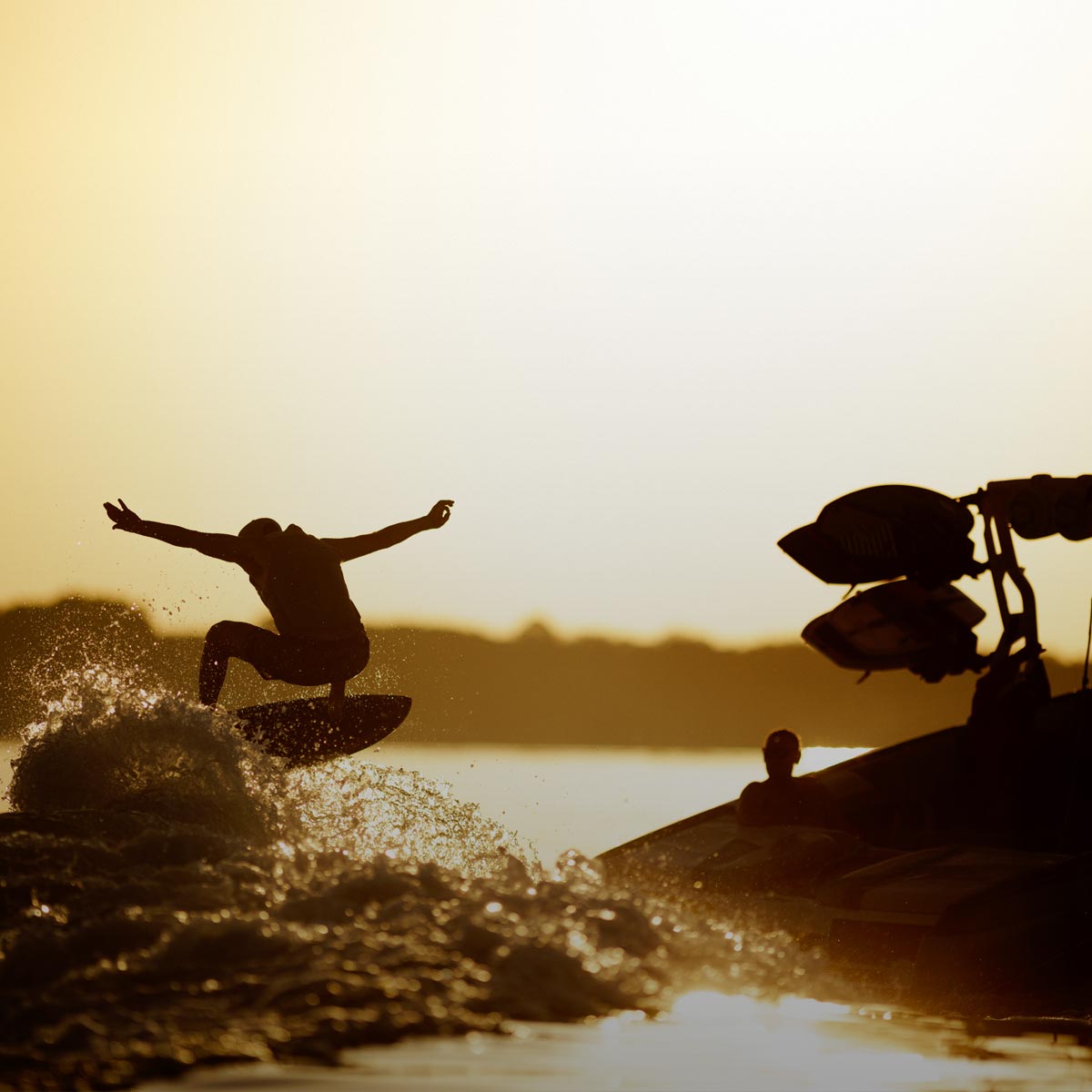 Silhouette of a wakesurfer catching air behind a boat at sunset with water spraying.
