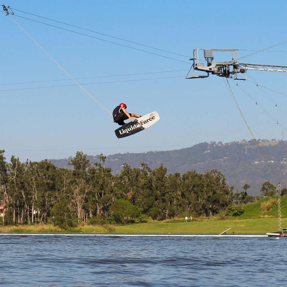 Busty D wakeboarding doing a huge jump on a lake with trees and mountains in the background. His white and black wakeboard showing the LiquidForce logo on the base.