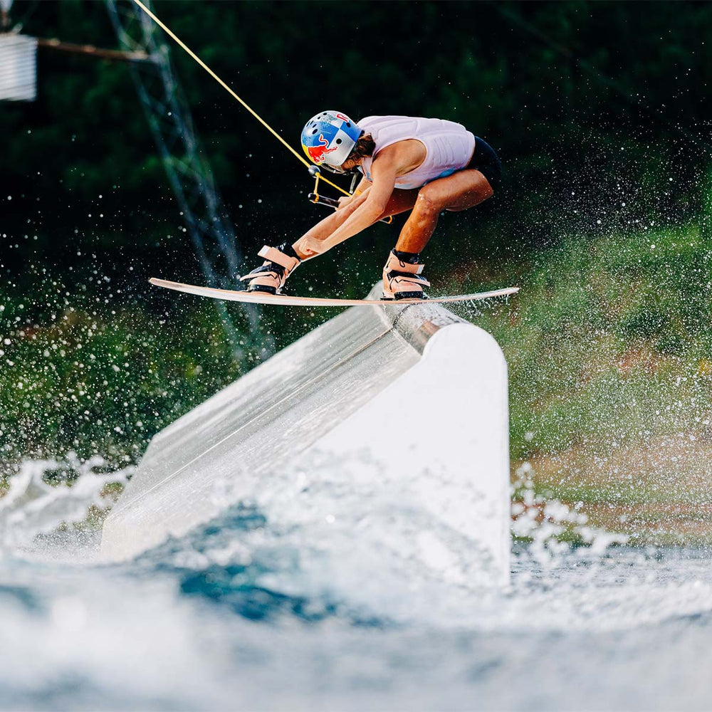 Claudia P doing a trick on a cable park feature on a wakeboard with pink bindings, a redbull helmet and pink life vest.