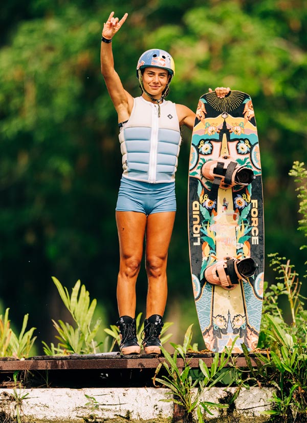 Claudia P holding a colorful wakeboard with pink bindings with a helmet and blue life vest, standing on a wooden dock with greenery in the background.