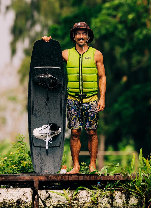 Man holding a black wakeboard with a green vest and helmet in a natural setting. Wearing a colorful swimsuit and a brown bucket hat.