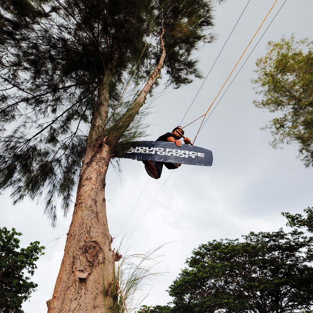 Felix G jumping over the camera on a wakeboard tapping a tree with his board. The base of the board shows black and white liquidForce logos.