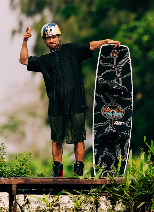 Felix G holding a black wakeboard standing on a dock with a natural background wearing a RedBull helmet.