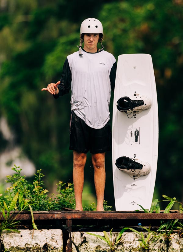 Gavin S holding a white wakeboard white bindings on a dock with a natural background. He is wearing a white helmet,  black swim suit and a white and black long sleeve shirt.
