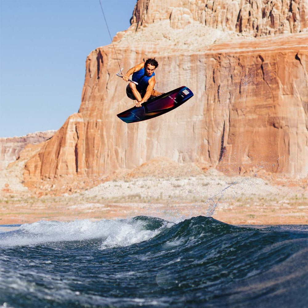 Guenther O jumping over a wake in front of a large rock formation. He is riding a colorful wakeboard and wearing a blue life vest.