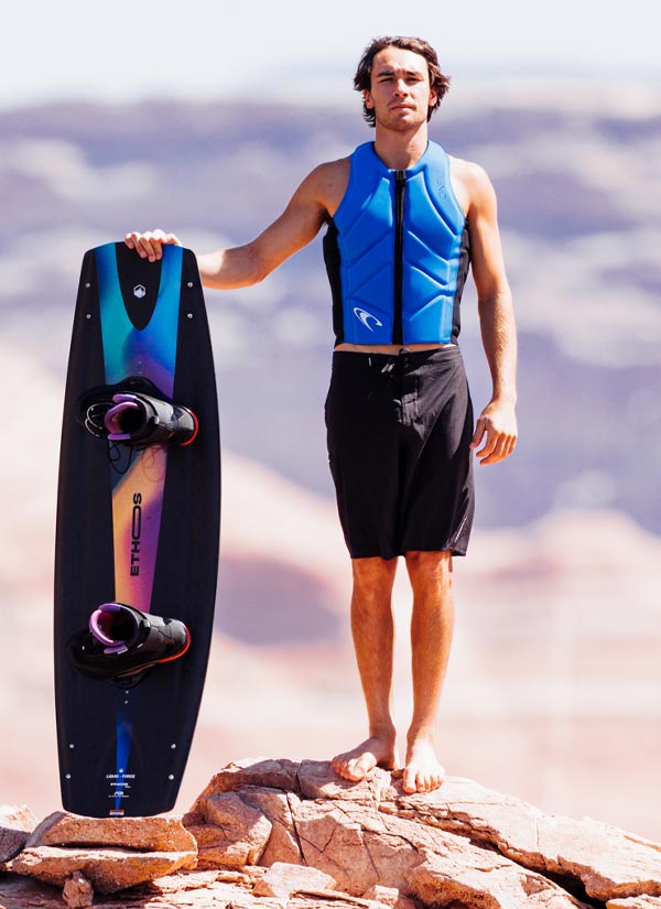 Guenther O holding a wakeboard with a colorful design, wearing a blue life vest and black shorts, standing on rocky terrain with a mountainous background.