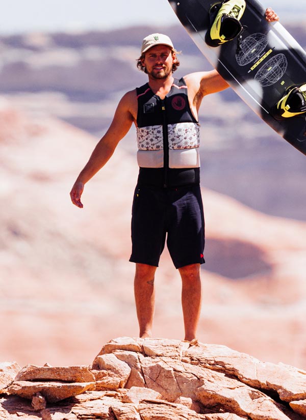 Harley C holding a black white and yellow wakeboard above his head on a rocky outcrop with a mountainous background wearing a white hat, black swimsuit and a colorful life vest.