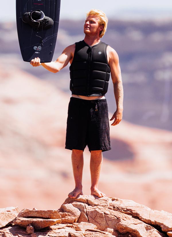 Luka K holding a wakeboard on a rocky outcrop with a scenic background. he is wearing a black life vest and black swimsuit.