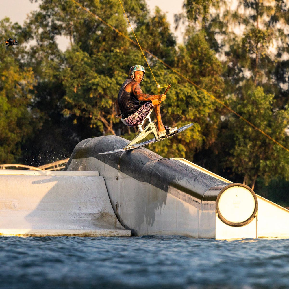 Wakeboarder grinding rail feature at cable park while holding handle, trees in background.
