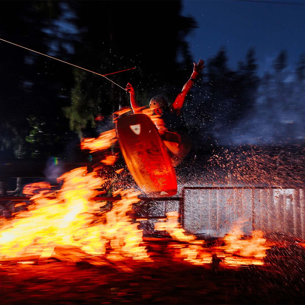 Matty M wakeboarding at night doing a trick over fire with a bright orange wakeboard and water splashes.