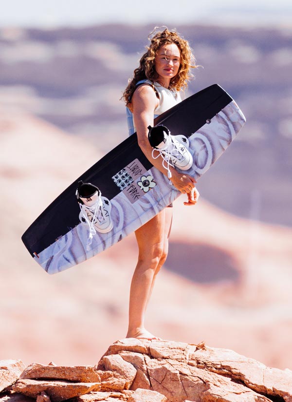 Meagan E holding a white and black wakeboard with a mountainous background.