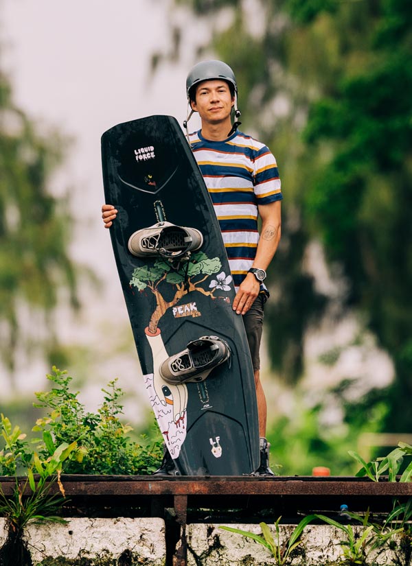 Nico V holding wakeboard with a nature-themed design standing on a dock wearing a striped shirt and a gray helmet.