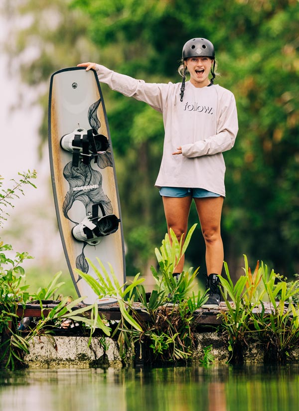 Rivers H holding a white and black wakeboard standing on a dock with a natural background. She is wearing a pink shirt, blue shorts and a black helmet.