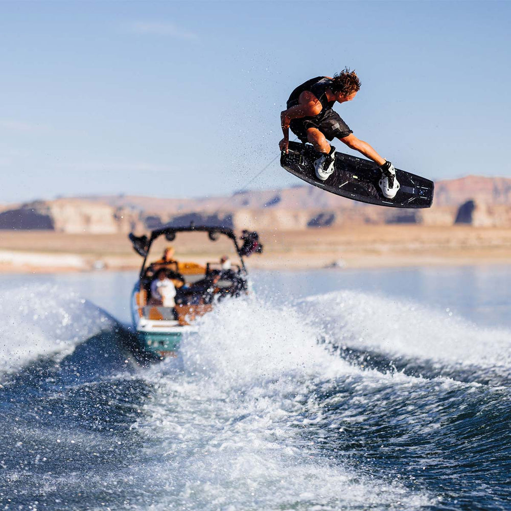 Sam B on a wakeboard performing a trick over a boat on water with mountains in the background. He is riding a black wakeboard with white bindings and wearing a black life vest with black swim suit.