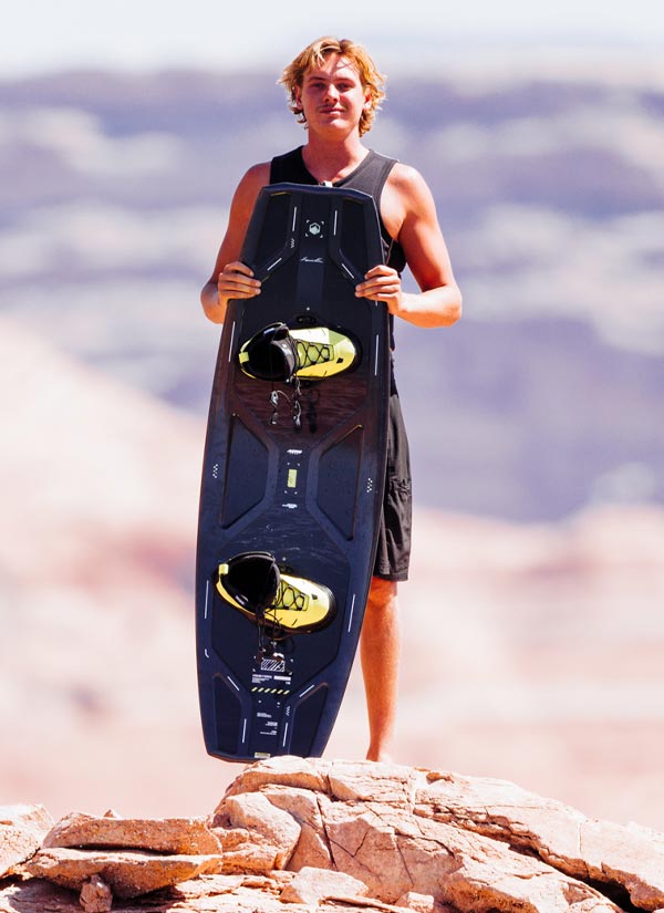 Sam B holding a blue wakeboard with yellow bindings on a rocky surface with a blurred natural background