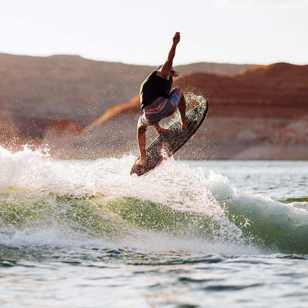 Person wake surfing on a wave with mountains in the background. He is wearing a colorful swimsuit and wearing a black life vest