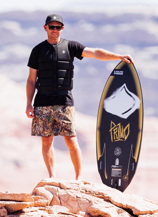 Tommy C holding a Primo wakesurf board on a rocky surface with a cloudy sky. He is wearing a black life vest and black hat with a camo swim suit.