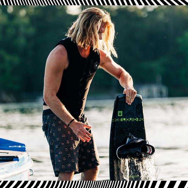 Man in vest holding dripping wakeboard beside a boat on a calm lake.