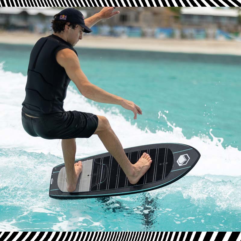 Man riding a black foil board barefoot while doing a trick on turquoise water.